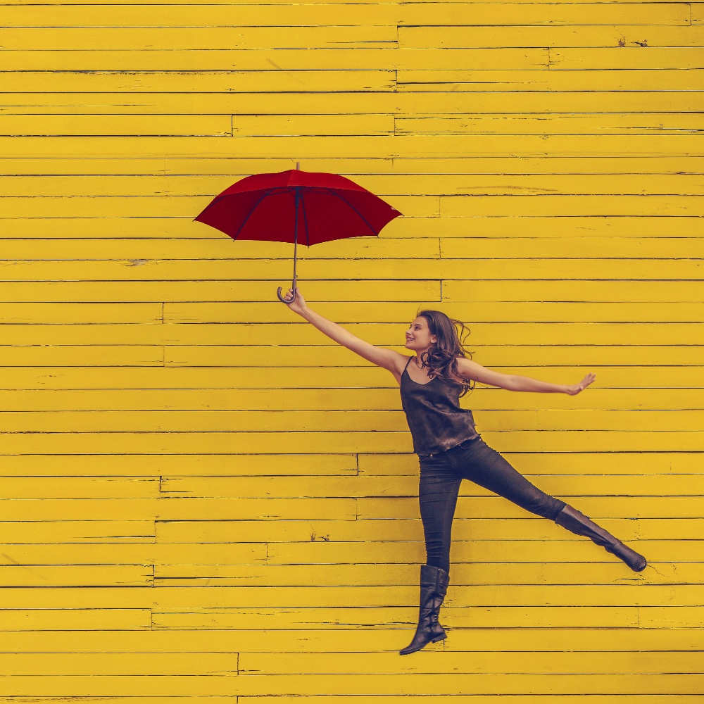 storytelling-lady A woman being lifted up into the air by a red umbrella, in front of an yellow wooden slat wall.